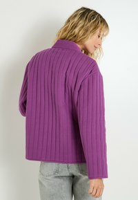 Veste matelassée violette avec un détail de côtes verticales, manches longues et coupe décontractée. Fabriquée à partir d'un tissu doux, elle présente un design sans col.