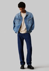 Giacca di jeans blu chiaro con due tasche sul petto, abbinata a una camicia beige e jeans blu scuro. Scarpe nere completano l'outfit.