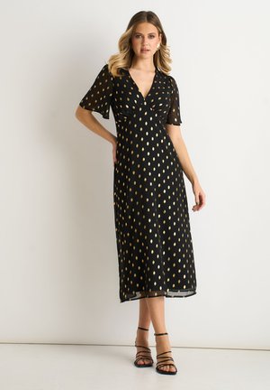 Woman in a black midi dress with gold polka dots, short flutter sleeves, V-neckline, and black strappy heels, standing against a white background.