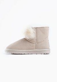 Bottine en suède beige avec un bout arrondi, accent en fausse fourrure douce en pompon, et une semelle en caoutchouc texturé avec un léger talon.