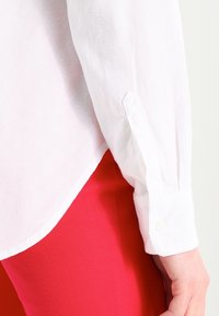 Chemise blanche à manches longues retroussées au poignet sur un pantalon rouge, main détendue et visible le long de la cuisse contre un fond neutre.