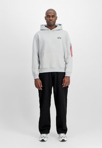 Sudadera de algodón gris con un bolsillo canguro, que presenta un logo y una etiqueta roja en la manga, combinada con pantalones negros y zapatillas plateadas.