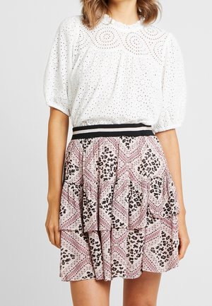 Kvinde iført en hvid bluse med puff-ærmer i eyelet-stof, kombineret med et lagdelt, pink og sort mønstret skørt med en sort og hvid taljebånd.