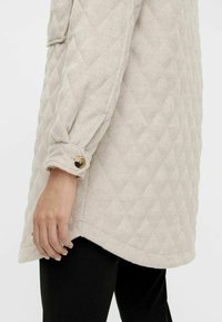 Chaqueta acolchada beige con una textura suave, que presenta un puño de manga plegable asegurado por un botón dorado, diseñada para un ajuste relajado.
