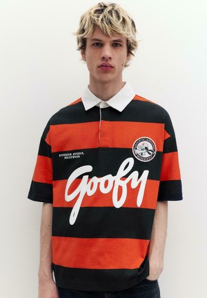 Młody mężczyzna o blond włosach, ubrany w czarną i pomarańczową polosshirt z paskami, z napisem "Goofy" i naszywką z kreskówkowym motywem na piersi.