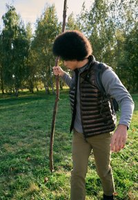 Mand går i naturen iført en mørkebrun dunvest, grå langærmet skjorte og khaki bukser, mens han holder en stok, omgivet af grønt. 