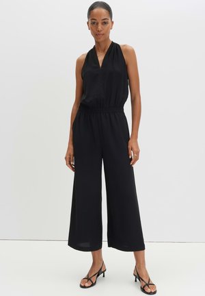 Vrouw draagt een mouwloze zwarte jumpsuit met wijde pijpen en V-hals, gecombineerd met zwarte hakken met bandjes, staand tegen een effen achtergrond.