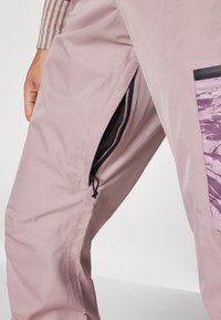 Helle rosa Hose aus glattem Stoff mit einer Reißverschlusstasche in schwarzer Hardware und einem Netzfutter für Belüftung. Abstraktes Musterelement an der Seite.