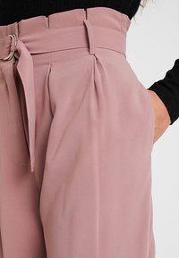 Light pink high-waisted trousers with a wide leg, featuring a belt and pleated details along the front, made from a smooth, draping fabric.