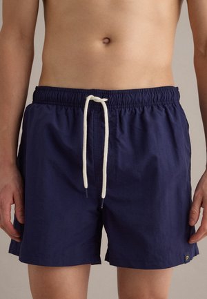 Male torso wearing navy blue elastic waist swim shorts with white drawstring, standing against a neutral background.