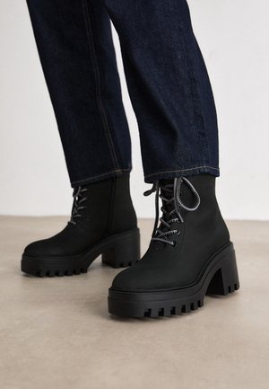 Svarta klumpiga platåstövlar med tjocka sulor med räfflat mönster och grå snören, båda tillsammans med mörkblå denimjeans, stående på ett ljust golv.