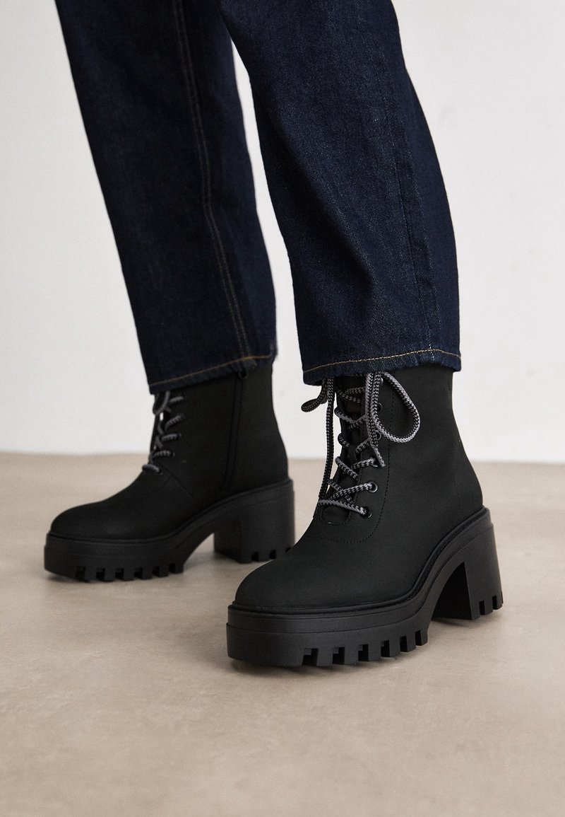 Czarne masywne botki na platformie z grubymi podeszwami o wyraźnym bieżniku i szarymi sznurówkami, noszone z ciemnoniebieskimi dżinsami, stojące na jasnej podłodze.