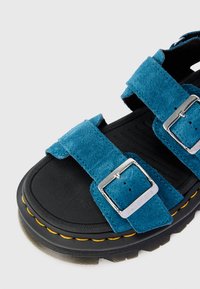 Sandașe din suede teal cu două curele ajustabile, catarame argintii și o talpă din cauciuc negru cu cusături galbene pe margine.