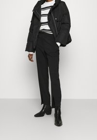 Schwarze Daunenjacke mit hohem Kragen, über einem weißen Pullover mit schwarzen Streifen getragen, kombiniert mit enganliegenden schwarzen Hosen und glänzenden schwarzen Stiefeln.