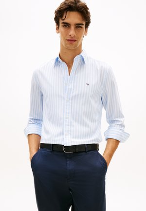 Fiatal férfi hullámos hajjal, világoskék csíkos ingben, sötétkék nadrágban és fekete övvel, kezei a zsebében.