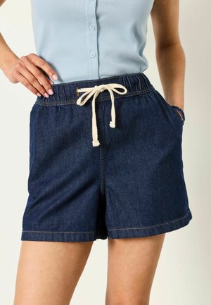 Žena nosí tmavomodré rifľové kraťasy s krémovým sťahovacím šnúrkou, svetlomodré bezrukávové košeľové tielko, ruka v vrecku a na boku.