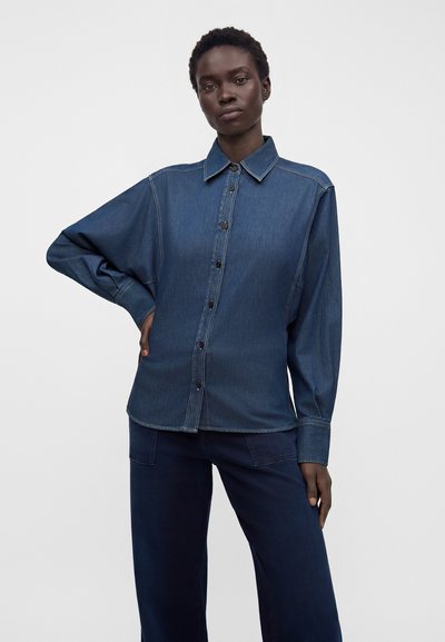 Modèle portant une chemise en denim bleu foncé à manches longues avec fermeture à boutons, associée à un pantalon bleu foncé assorti, debout sur fond blanc.
