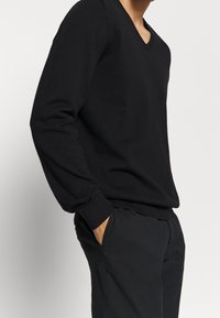 Schwarzer V-Ausschnitt-Pullover mit langen Ärmeln, gerippten Bündchen und Saum, getragen zu dunkler Hose. Der Stoff wirkt glatt und leicht.