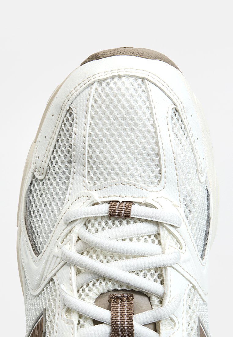 Zapatilla de malla blanca con una superficie texturizada, acentos en marrón, punta redondeada y cordones planos. Suela reforzada y acolchada para mayor comodidad.