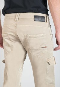 Beige cargo broeken met meerdere zakken, met een leren patch op de tailleband. Zachte textuur en gestructureerde, taps toelopende pasvorm.