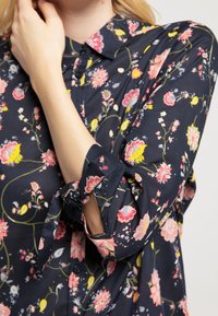 Blusa azul marino con estampado floral en rosa, amarillo y verde. Presenta un cierre frontal con botones, cuello y detalles de lazo en los puños.