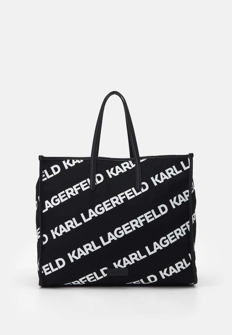 Svart tote-väska med vit "KARL LAGERFELD" text tryckt diagonalt. Har långa svarta handtag och en slitstark tygkonstruktion.