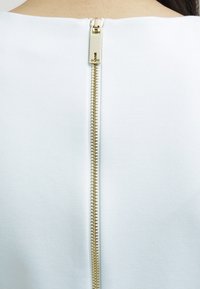 White fabric with a smooth texture, featuring a gold zipper at the back, extending from the neckline to the lower back.