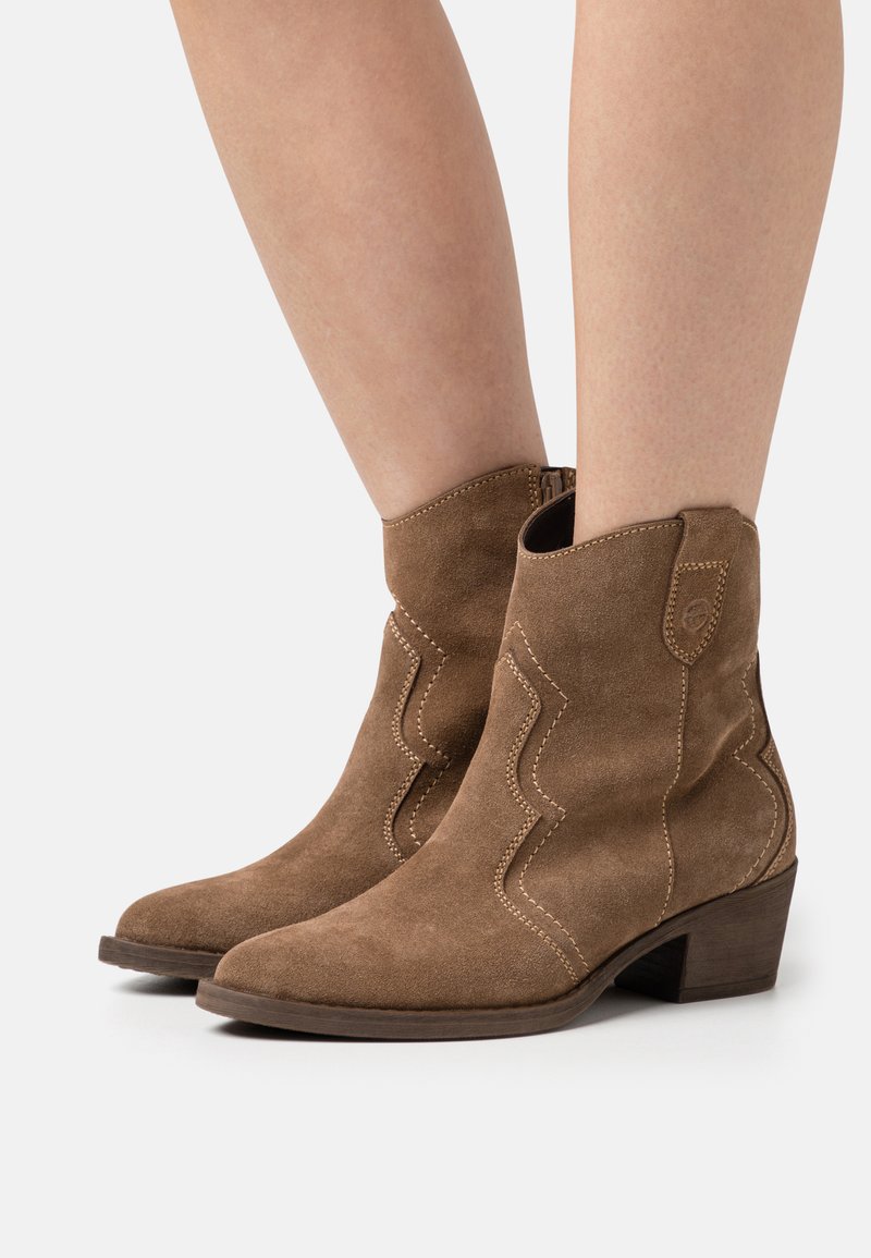 Botas de tobillo de ante marrón con un diseño western, que presentan costuras curvadas y un tacón bajo. La textura es suave y ligeramente desgastada.