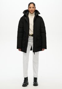 Czarna ocieplana parka z wysokim kołnierzem, dwoma przednimi kieszeniami i zapięciem na zamek. Nosi się ją na kremowym golfie i zestawia z białymi spodniami oraz czarnymi butami.