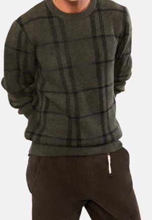 Uomo con maglione a quadri verde scuro con linee nere e pantaloni di velluto a coste marrone scuro, mani dietro la schiena su sfondo bianco.