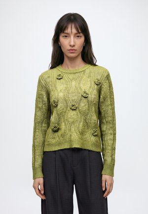 Vrouw draagt een groene kabelgebreide trui met kleine bloemdetails, gecombineerd met zwarte broek met wijde pijpen, staand tegen een effen achtergrond.