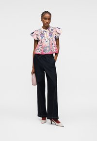Femme portant un chemisier rose à volants avec motif floral, un pantalon noir à jambes larges, des talons blancs et tenant un petit sac à main rose, posant devant un fond blanc.
