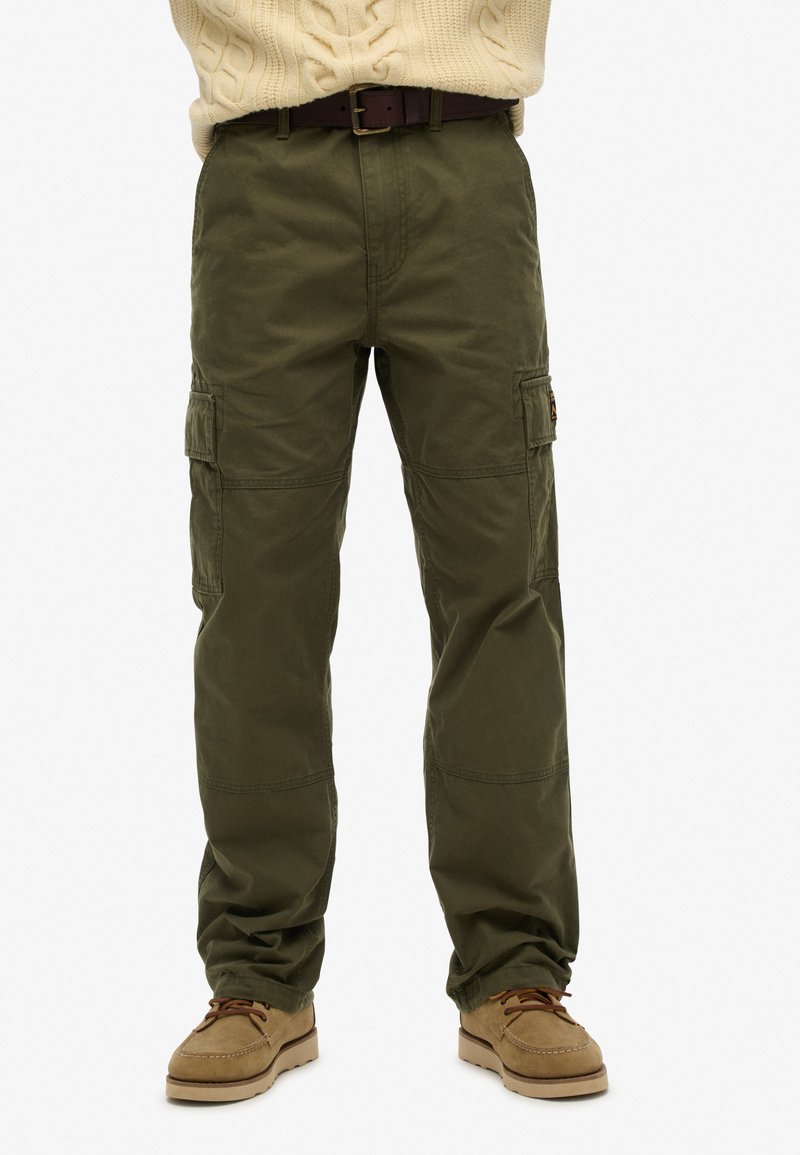 Olijfgroene cargo-broek met zijzakken, een wijde pasvorm en een zachte textuur. Gepaard met een lichtbeige kabelgebreide trui en bruine schoenen.