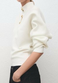 Femme portant un pull blanc côtelé à boutons dorés et un pantalon noir, posant avec une main dans la poche devant un mur uni.