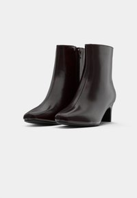 Pár tmavě hnědých kožených kotníkových bot s nízkými podpatky a bočními zipy, vyfocených na jednoduchém bílém pozadí.