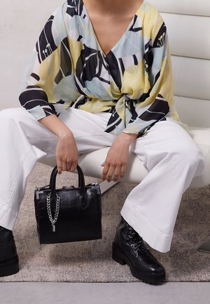 Osoba ubrana w żółtą, niebieską i czarną bluzkę w wzory, białe szerokie spodnie i czarne buty, trzymająca czarną torebkę z srebrnym łańcuszkiem.