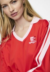 Rotes Sporttrikot mit pinkem Kragen, das ein Logo mit einem Fußball und einem Karomuster zeigt, sowie wei�ßen Streifen an den Ärmel.