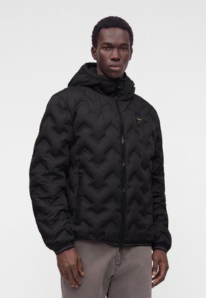 Jeune homme portant une veste matelassée noire à capuche, debout devant un fond clair uni, les mains détendues sur les côtés.