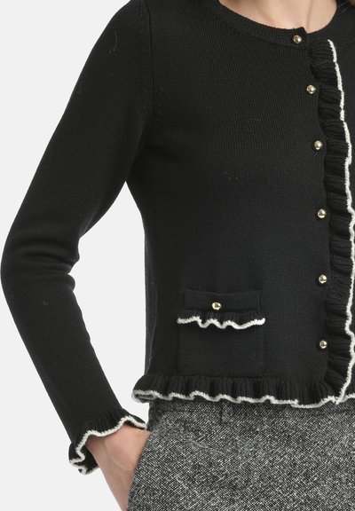 Melna iestrēdzusi kardigāna jaka ar baltu frillē joņu, zeltītiem pogām un teksturētu kabatu ar frillēm. Garas piedurknes ar rūtainu malu.