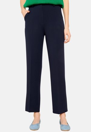 Frau trägt marineblaue, gerade geschnittene Hosen, grünes Oberteil und hellblaue Ballerinas, steht mit einer Hand in der Tasche.