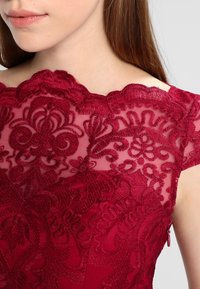 Burgundská krajková blůza s výšivkami květinových vzorů, s vlnitým výstřihem a průhlednou vrstvou; obsahuje detail bočního zipu.