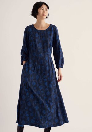 Woman with short black hair wearing a long blue dress with floral patterns and black ankle boots, standing against a plain light background.