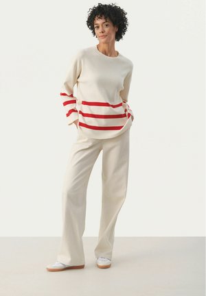 Suéter color crema con rayas rojas horizontales y dos botones en los lados, combinado con pantalones de pierna ancha beige claro y zapatillas blancas.