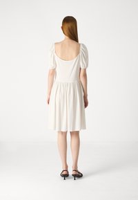 Robe blanche avec de courtes manches bouffantes et un dos plongeant. Jupe évasée avec une taille froncée. Tissu lisse, longueur au genou, portée avec des chaussures noires.