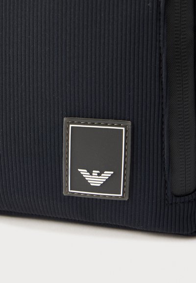 Κοντινή λήψη υφάσματος σε ναυτικό μπλε με υφή και τετράγωνο μαύρο-άσπρο λογότυπο με στυλιζαρισμένη εικόνα αετού στη γωνία.