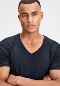 Navyblaues V-Ausschnitt-T-Shirt aus weichem Stoff, taillierte Passform, mit kurzen Ärmeln und dezenten Steppdetails. Das Modell posiert mit verschränkten Armen.