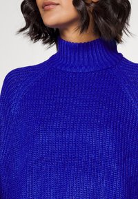 Lila stickad polotröja med ribbad textur och raglansnitt, som har en hög krage och en lös passform.