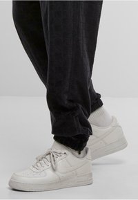 White Nike Air sneakers worn with black textured pants and white socks, standing on a plain light gray floor.