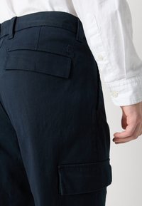 Sötétkék cargo nadrág texturált felülettel, oldalsó zsebekkel és egy logóval ellátott hátsó zsebbel rendelkezik. Fehér inggel viselik.