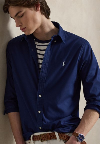 Νεαρός άνδρας που φοράει μπλε πουκάμισο με κουμπιά πάνω από ριγέ μπλουζάκι, μπεζ παντελόνι με πλεκτή καφέ ζώνη και μπλε ρολόι χειρός.